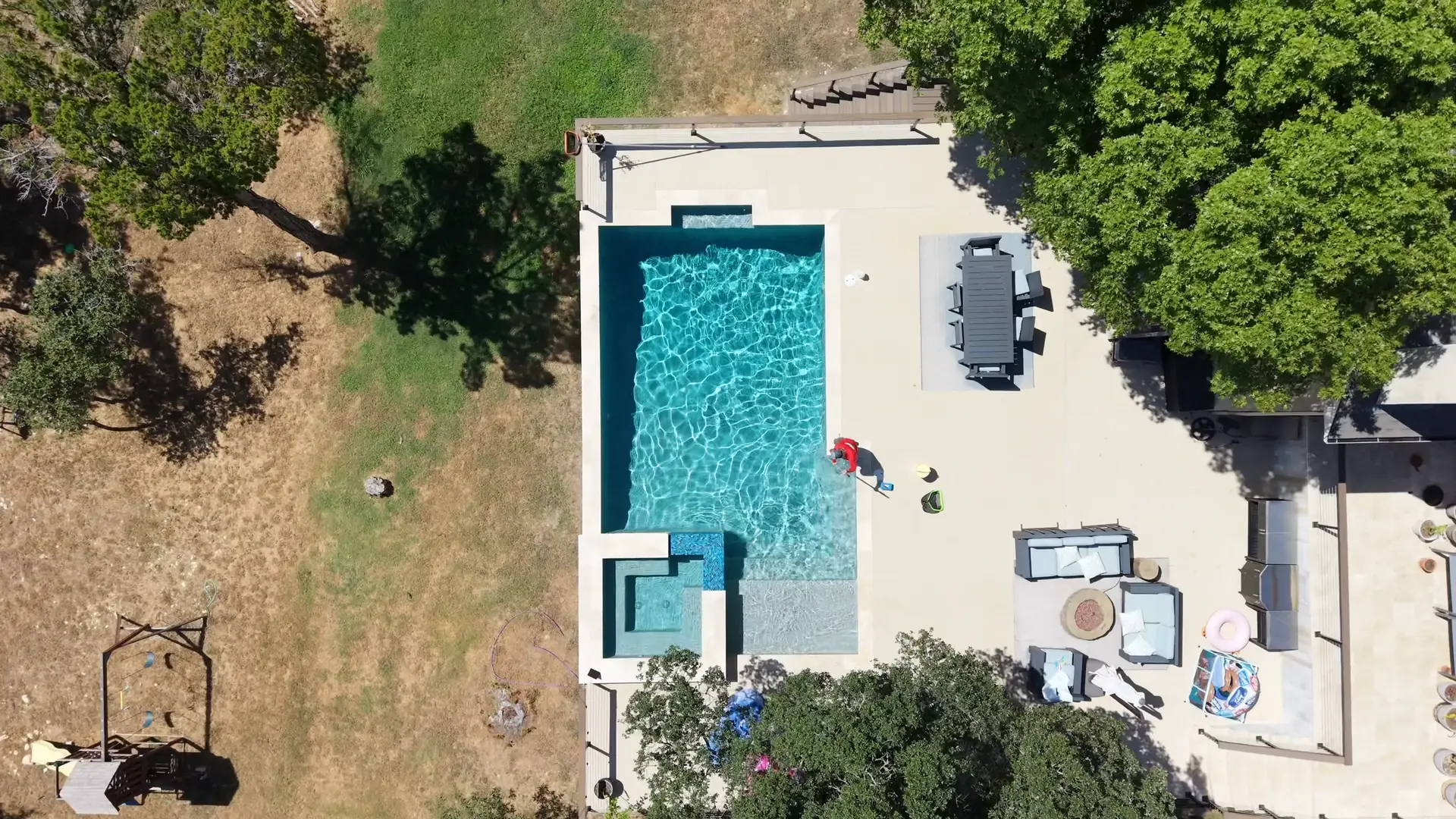 Aerial views over Austin hill-country back gardens — rectangular pools and spas in pale stone terraces, live oaks and sun-bleached grass around them — cut with a technician in a red Sullivan’s shirt working at the water’s edge with a net on a long pole.