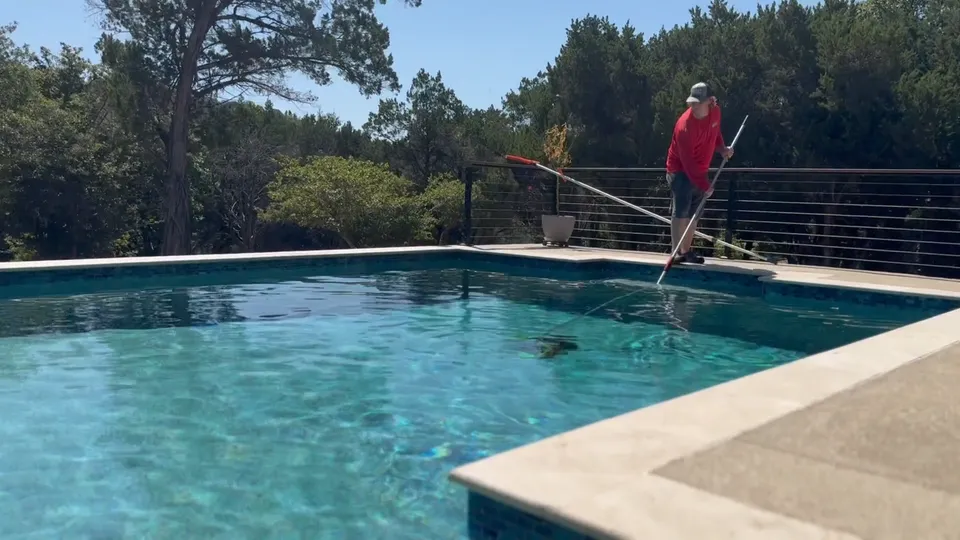 A Sullivan’s technician in a red shirt skimming a pool with a long pole, beside a cable-rail fence and live oaks