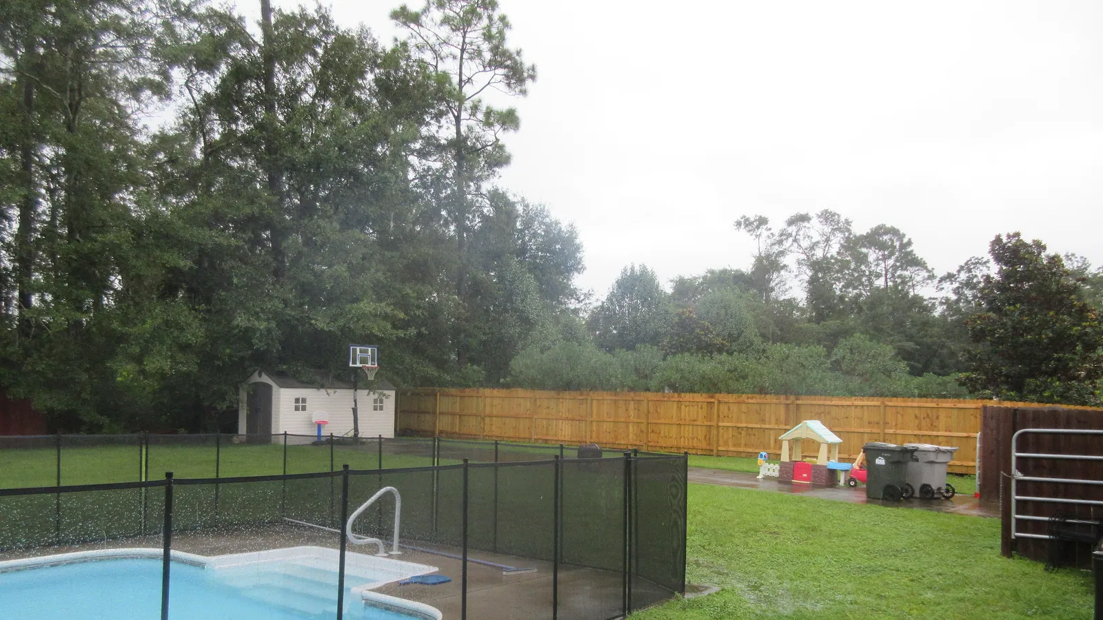 A backyard pool behind black mesh safety fencing, with a timber privacy fence, a small garden shed and children’s play equipment on the grass beyond it.