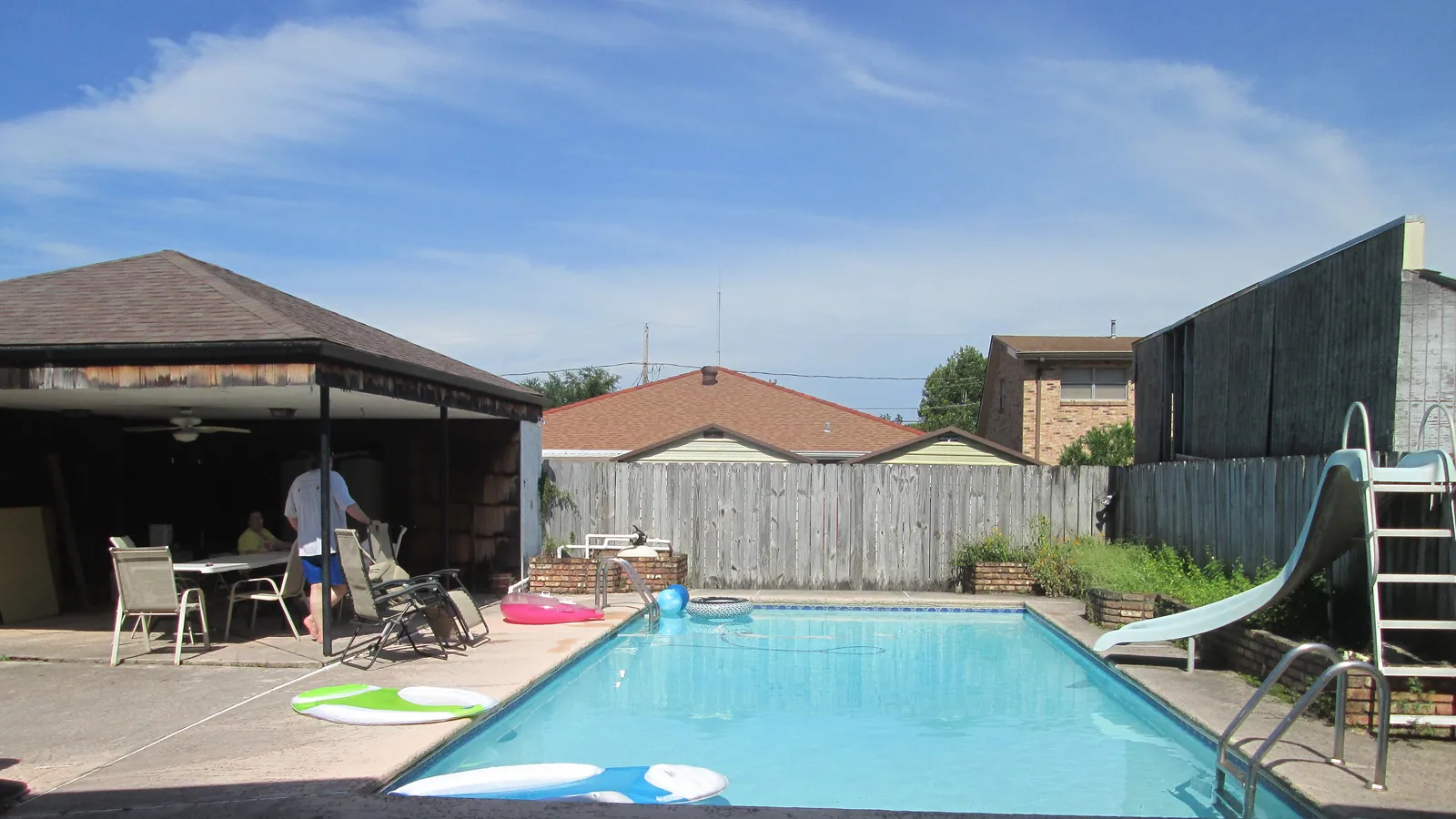 A backyard pool with a white curved slide at the far end, floats lying on the paved surround, a timber fence behind and, under the carport to one side, patio chairs and a person standing with their back to the camera.