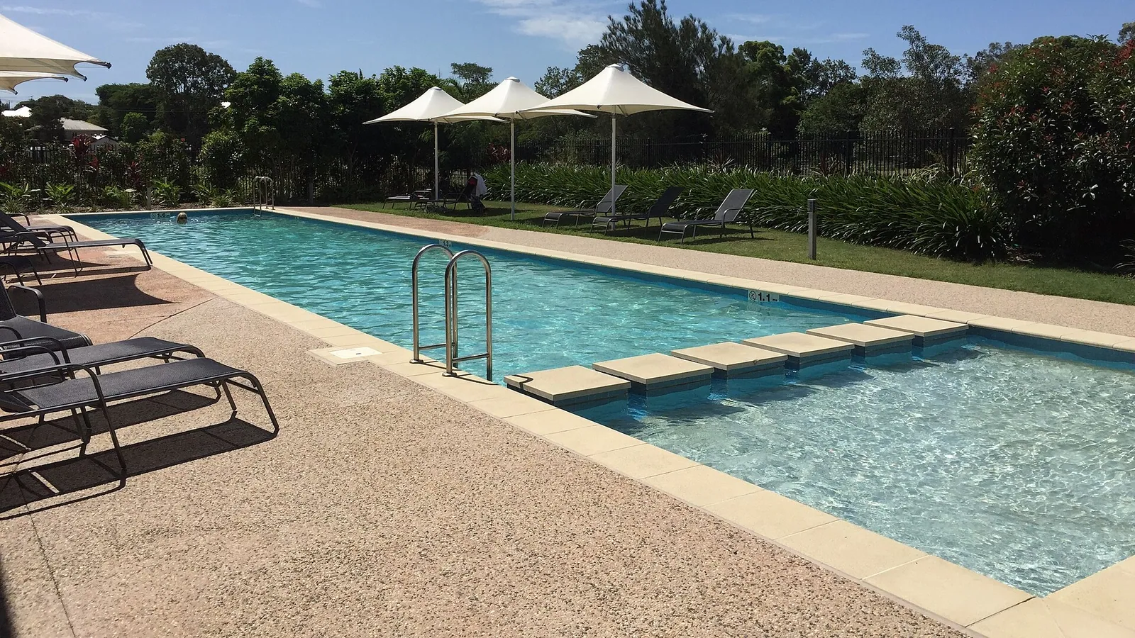 A long pool with a stainless steel handrail at the near steps, loungers on the paved surround and white parasols along the far side.