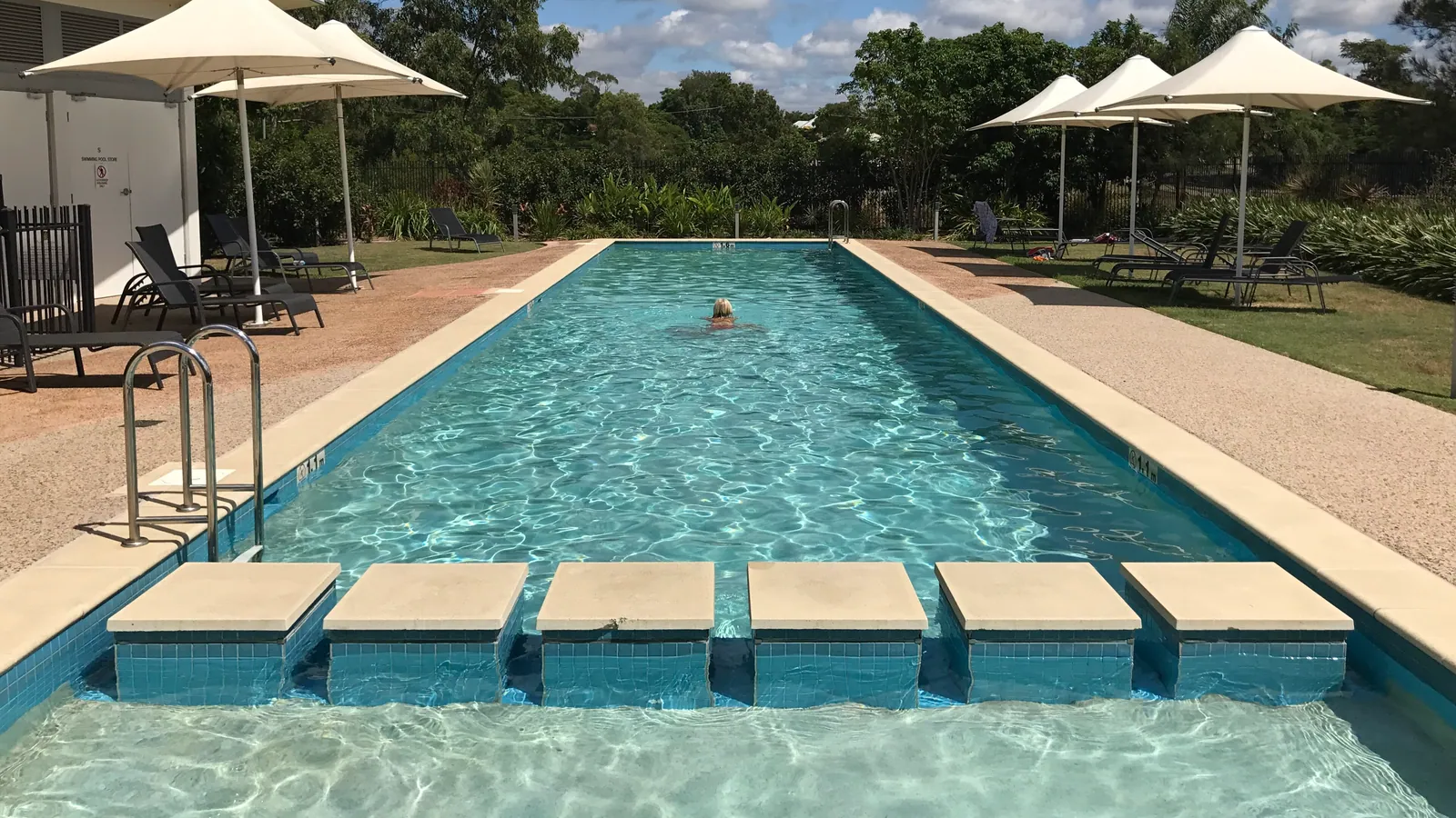 A long pool with square stone blocks set across the near end as a walkway, one distant swimmer in the far half, and white parasols and loungers on the surround beyond.